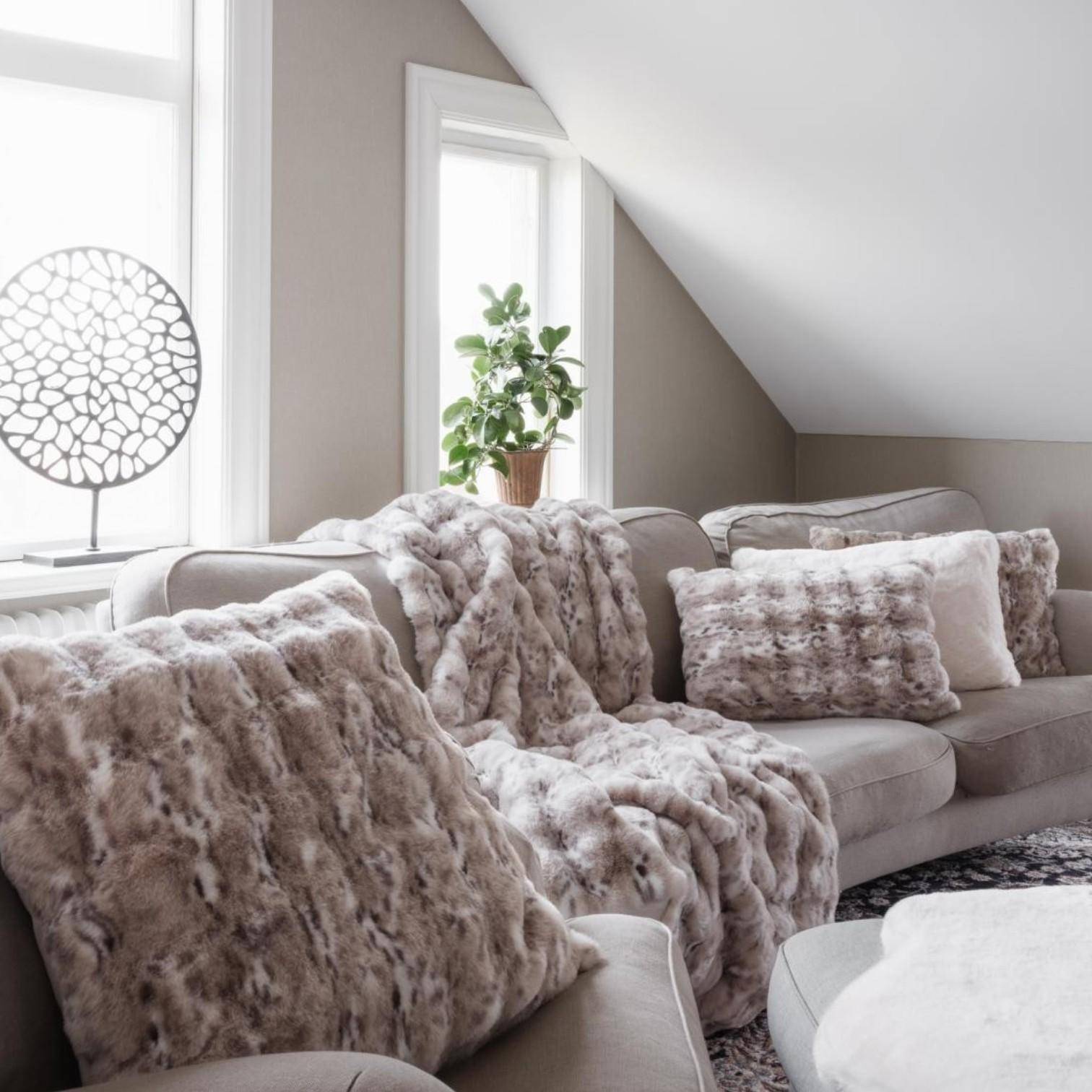 Skinnwille Plaid Nika Snow Leopard mit passenden Dekokissen als stilvolle Kuscheldecke auf einem modernen Sofa im gemütlichen Wohnzimmer.