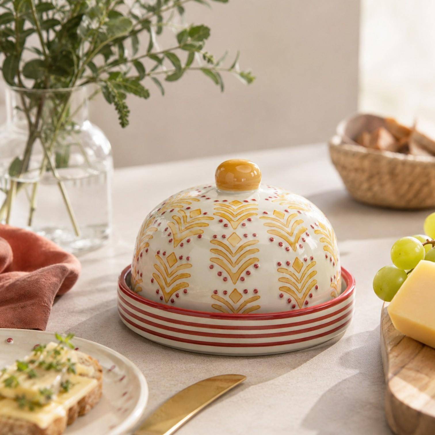 Handgefertigte Tranquillo Käseglocke Oriental aus Steinzeug mit gelb-rotem Muster in einem mediterranen Ambiente mit Käse, Trauben und natürlichen Dekoelementen.