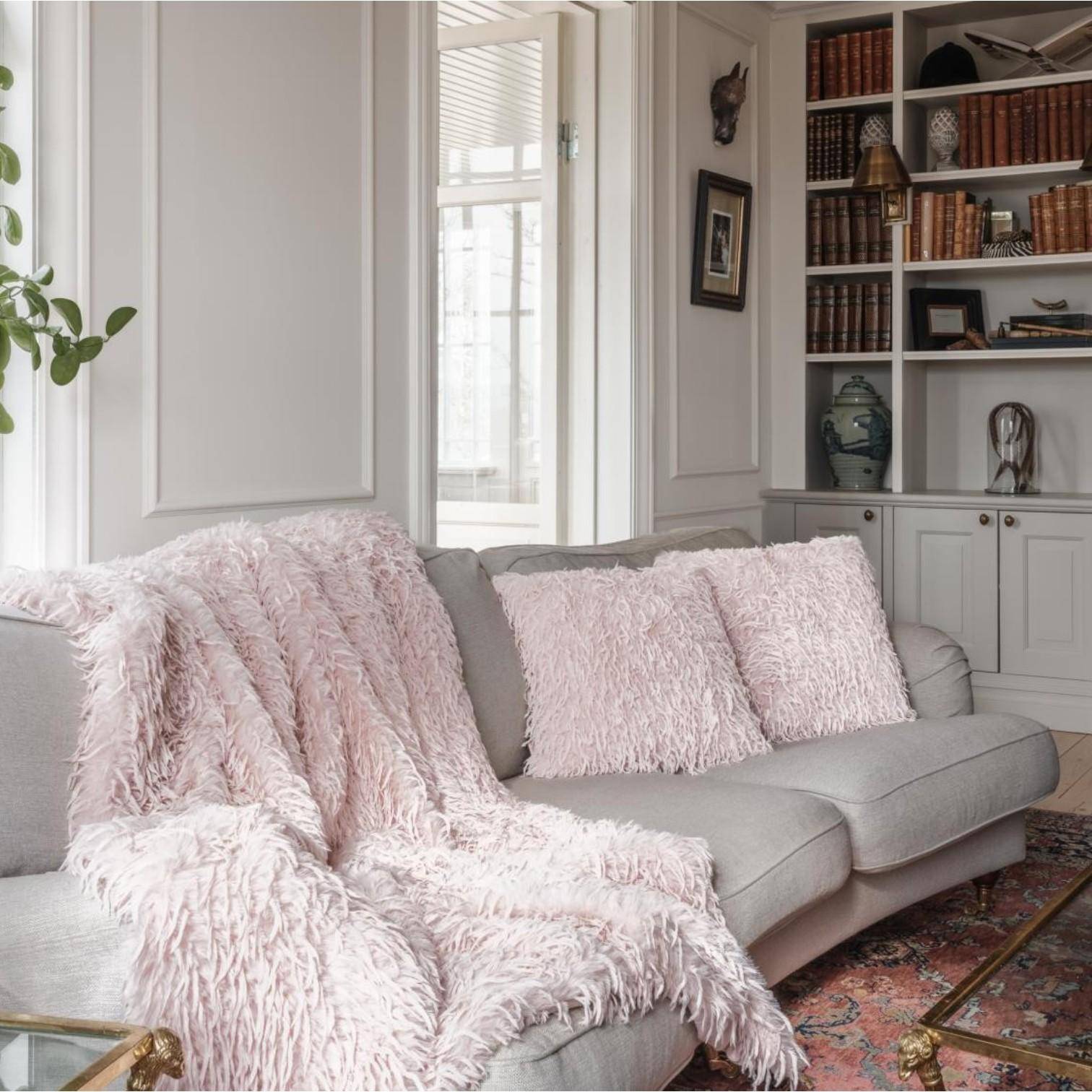 Skinnwille Dekokissen Fay in Pale Pink 45x45 cm mit federähnlicher Vorderseite auf einem hellen Sofa, kombiniert mit einer kuscheligen Kunstfelldecke in stilvollem Wohnambiente.